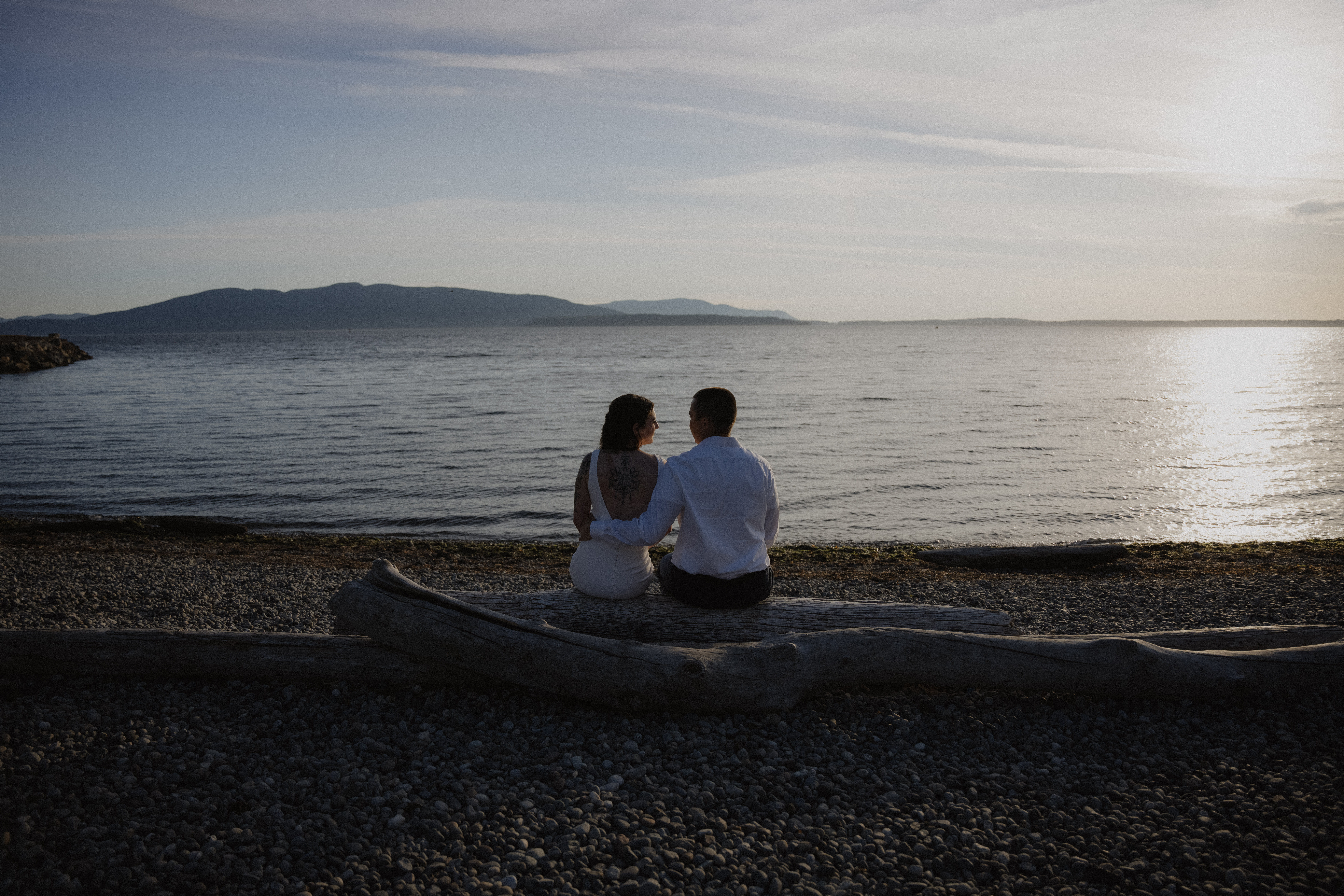 bride and groom sit on log staring out at the ocean during bellingham engagement shoot